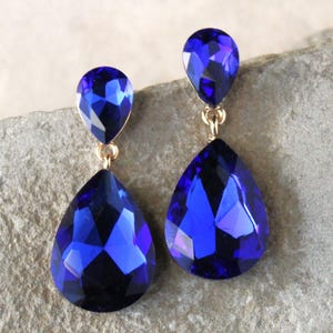 Op de afbeelding: Een paar oorhangers met twee druppelvormige blauwe kristallen aan elke oorhanger. De kristallen zijn facetgeslepen en hebben een gouden setting.