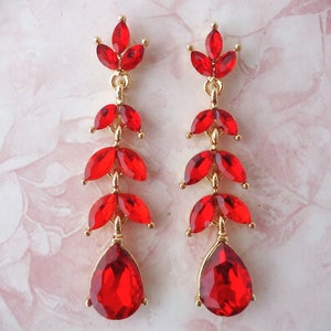 May include: A pair of long, dangling earrings with a gold-tone metal setting. Each earring features a series of red, leaf-shaped gemstones, culminating in a large, teardrop-shaped gem at the bottom. The earrings are designed for pierced ears.