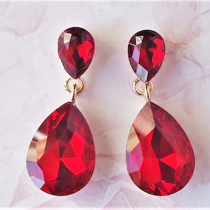 May include: A pair of red teardrop-shaped dangle earrings with gold accents. The earrings feature two red crystal drops, one smaller than the other.