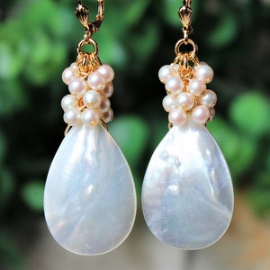 May include: A pair of drop earrings featuring large, white, teardrop-shaped mother-of-pearl pendants suspended from clusters of small, round, pale pink pearls. The components are connected by gold-toned metal rings and lever-back earring hooks.
