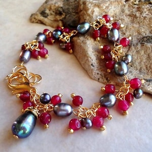 May include: A charm bracelet featuring gold-tone metal links adorned with clusters of dark grey oval pearls and small, round, vibrant red beads. The bracelet includes a gold-tone lobster claw clasp and a larger, teardrop-shaped grey pearl charm.