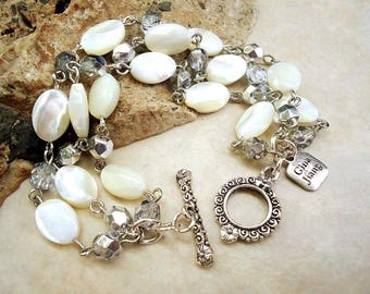 May include: A multi-strand bracelet featuring oval white mother-of-pearl beads interspersed with faceted translucent gray beads on a silver-tone chain. The bracelet includes a decorative toggle clasp and a square metal tag engraved with Gina Jiang.
