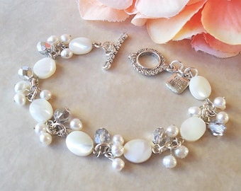 May include: A delicate bracelet featuring white, oval-shaped mother-of-pearl beads, small white pearls, and faceted silver beads. The bracelet has a silver toggle clasp and a small tag that reads "Gina Jiang".