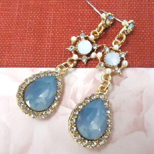 May include: A pair of gold tone dangle earrings with blue teardrop shaped stones surrounded by clear rhinestones. The earrings also feature a white stone surrounded by clear rhinestones and small white pearls.