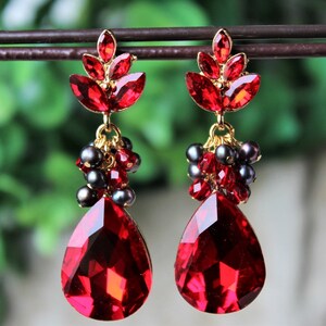 May include: A pair of dangle earrings with red teardrop-shaped crystals and black beads. The earrings are gold-toned and have a cluster of smaller red crystals at the top.