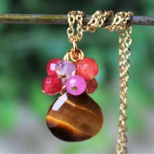 May include: A gold-colored necklace featuring a pendant with a teardrop-shaped tiger's eye stone. The pendant is accented with a cluster of small, colorful beads in shades of red, pink, and clear. The necklace hangs on a delicate chain.