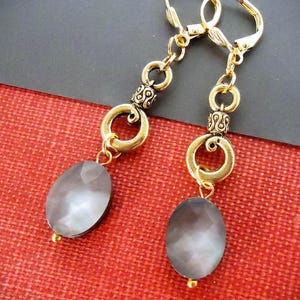 May include: A pair of dangle earrings featuring gold-tone metal leverback closures, decorative textured rings, and oval-shaped faceted gray stones. The earrings have a metallic finish and a dangling design that hangs from the top metal loop components.