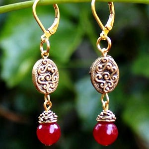 May include: A pair of gold-colored earrings with an oval, ornate design and a red, round bead. The earrings hang from a gold lever-back clasp. The background is a blurred green.