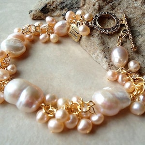 May include: A pearl bracelet with large, irregular pearls and smaller, clustered pearls in cream and peach hues. The bracelet has gold-coloured chain links and a decorative clasp. A small gold tag reads "Gina Jiang".