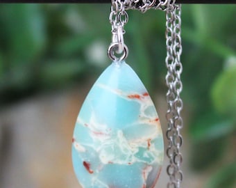 May include: A silver necklace with a teardrop-shaped pendant. The pendant is a light blue stone with red and white marbling. The chain is silver. The background is a blurred green.