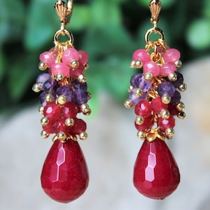 May include: Dangling earrings with a gold-tone metal setting. Each earring features clusters of pink, purple, and red beads, culminating in a faceted, teardrop-shaped red gemstone. The earrings have a lever-back closure.