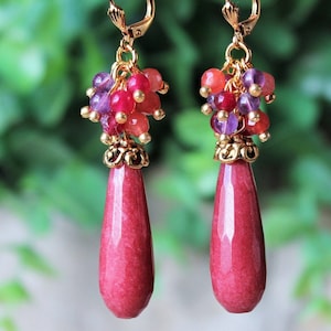 May include: A pair of dangle earrings with red and purple gemstone beads and gold accents. The earrings feature a teardrop-shaped red stone.