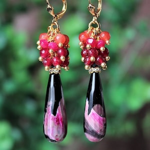 May include: A pair of dangle earrings with gold-colored leverback closures. Each earring features a cluster of red and orange beads above a teardrop-shaped pendant. The pendants are black with pink and white swirls.