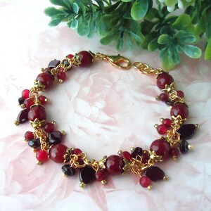 May include: A charm bracelet featuring deep red and dark burgundy beads in various shapes and sizes, accented with gold-toned metal caps and links. The bracelet includes a gold-toned lobster claw clasp and a short extender chain for adjustable length.