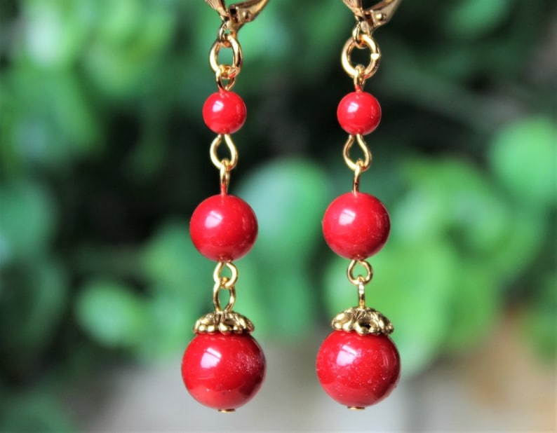 May include: A pair of red bead earrings with gold accents. The earrings are made with three round beads and a gold hook.