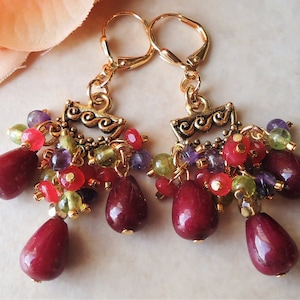 以下が含まれることがあります： 赤、緑、紫、金のビーズがついた、ゴールドのドロップイヤリングのペア。イヤリングの上部には、装飾的なゴールドチャームが付いています。