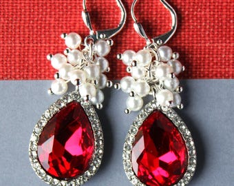 May include: A pair of drop earrings featuring a large, pear-shaped red crystal surrounded by a halo of small clear rhinestones. Above the crystal, a cluster of small white faux pearls is attached to a silver-toned metal lever-back earring hook.