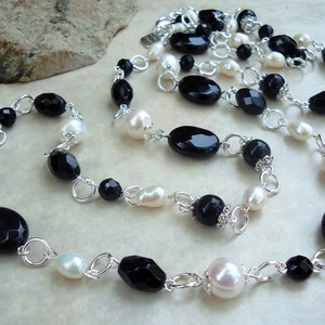 Puede incluir: Un collar largo con cuentas alternas de ónix negro y perlas blancas, conectadas por eslabones plateados. El collar presenta una mezcla de cuentas ovaladas y redondas, creando un diseño clásico y elegante.