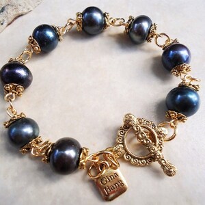 May include: A gold-toned bracelet featuring dark blue pearls. The bracelet has ornate gold connectors and a decorative clasp. A small gold tag with the text "Gina Jiang" hangs from the clasp.
