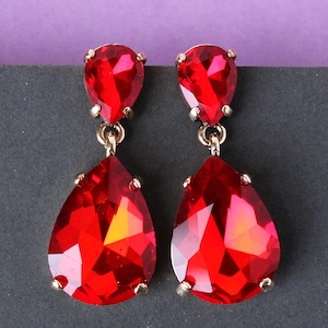 May include: A pair of red teardrop earrings with gold-coloured metal accents. Each earring features a smaller, rounded gem at the top and a larger, pear-shaped gem suspended below. The gems have a faceted appearance, reflecting light.