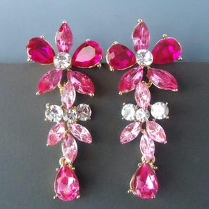 May include: A pair of pink and clear crystal flower dangle earrings. The earrings feature a large flower with pink and clear crystals, followed by a smaller flower with clear crystals, and a teardrop-shaped pink crystal.