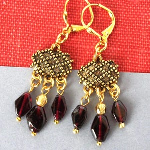 May include: A pair of gold-tone dangle earrings featuring a textured, diamond-patterned metal top piece. Each earring has three dangling beads, including two dark red faceted beads and one smaller, translucent yellow bead suspended by gold links.