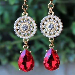 May include: Gold dangle earrings with a red teardrop-shaped stone and a circular design with white pearls and rhinestones.