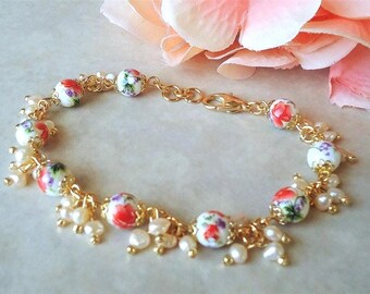 May include: A gold-toned bracelet featuring floral-patterned beads in red, white, and purple, interspersed with small white pearls. The bracelet is connected by a delicate gold chain and has a clasp closure. The design is feminine and elegant.