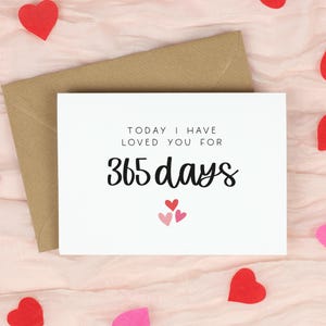 May include: A white card with the text "TODAY I HAVE LOVED YOU FOR 365 days" and three small hearts. A brown envelope is behind the card. Red and pink heart-shaped confetti is scattered on a pink fabric background.