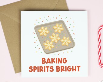 May include: A square greeting card with the text "BAKING SPIRITS BRIGHT" features a baking sheet with snowflake cookies. A brown envelope, snowflakes, and a candy cane heart are on a pink background.