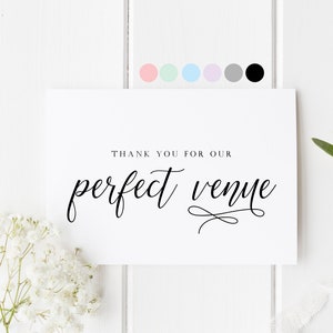 May include: White card with black calligraphy that reads "Thank you for our perfect venue". The card is on a white wood background with white flowers and a green sprig.