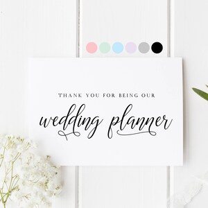 May include: A white card with the text "THANK YOU FOR BEING OUR wedding planner" in elegant black script. The card is surrounded by white flowers, greenery, and a white ribbon, with a palette of pastel color swatches.