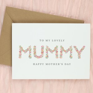 May include: A white greeting card with the words "TO MY LOVELY MUMMY HAPPY MOTHER'S DAY". The word "MUMMY" is designed with pink and peach flowers. A brown envelope is behind the card.