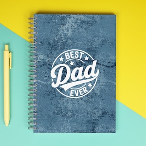 May include: A spiral-bound notebook with a blue textured cover and the words "Best Dad Ever" in a white circular design. A yellow pen is positioned to the left of the notebook against a teal and yellow background.