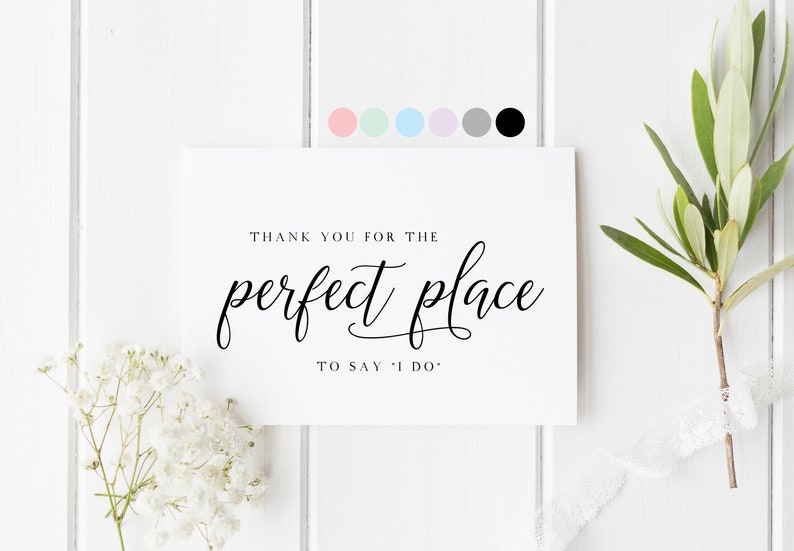 Op de afbeelding: Een witte kaart met de tekst "THANK YOU FOR THE perfect place TO SAY 'I DO'" in elegant zwart schrift. De kaart is versierd met witte bloemen en groen, wat duidt op een bruiloftsthema.