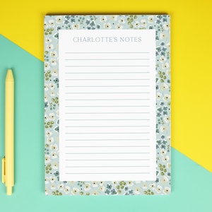 May include: A notepad with the text "CHARLOTTE'S NOTES" on a white background, with a floral pattern in blue, green, and white. A yellow pen lies on a turquoise and yellow background.