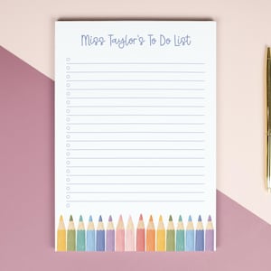 May include: A white rectangular notepad with the text Miss Taylor's To Do List printed in blue at the top, featuring horizontal lines with circular bullet points and a row of colourful pencil illustrations at the bottom, paired with a gold pen.