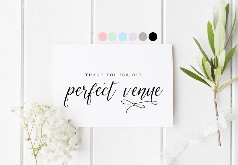 May include: White card with black calligraphy that reads "Thank you for our perfect venue". The card is on a white wood background with white flowers and a green sprig.