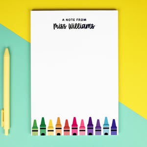 May include: A white rectangular notepad featuring the black printed text A NOTE FROM Miss Williams at the top and a row of colourful crayon illustrations along the bottom edge, accompanied by a single yellow retractable ballpoint pen.