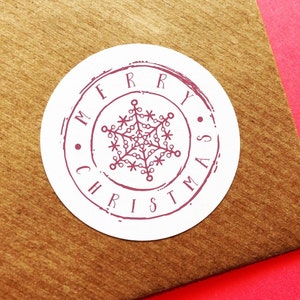 May include: A white, circular sticker with a burgundy design and the words "MERRY CHRISTMAS" encircling a snowflake. The sticker is on a brown surface with a red background.