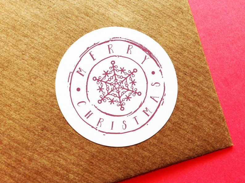 May include: A white, circular sticker with a burgundy design and the words "MERRY CHRISTMAS" encircling a snowflake. The sticker is on a brown surface with a red background.