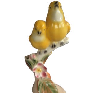 May include: A ceramic figurine of two yellow birds perched on a branch with pink flowers and green leaves. The birds have white bellies and are looking upwards. The base is a light peach color.