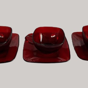 May include: Three ruby red glass teacups and saucers. Each cup has a square shape with a handle, resting on a matching square saucer. The set is made of transparent glass, reflecting light.