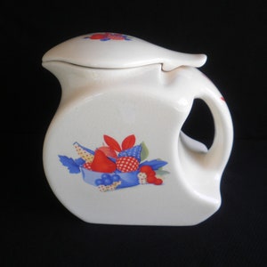 May include: White ceramic pitcher with a lid and a fruit design. The pitcher has a handle and a spout. The fruit design features red, blue, and yellow fruits with green leaves.