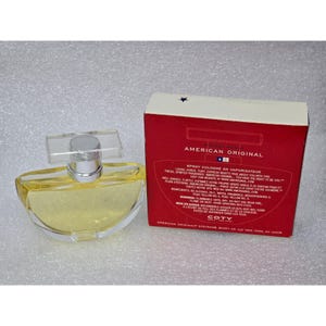 May include: A clear glass perfume bottle with a silver cap and a rectangular clear top, next to a red box with the words "American Original" in white. The bottle contains a yellow liquid.