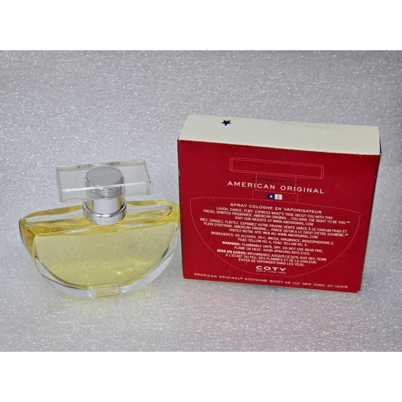 May include: A clear glass perfume bottle with a silver cap and a rectangular clear top, next to a red box with the words "American Original" in white. The bottle contains a yellow liquid.