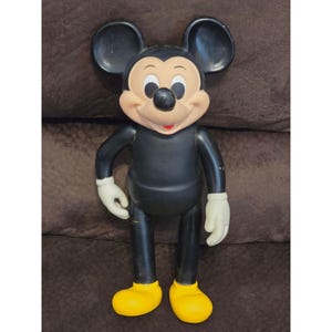 May include: A vintage Mickey Mouse figurine. The figure has a black body, large black ears, a tan face, white gloves, and yellow shoes. The figure is standing upright with arms at its sides.