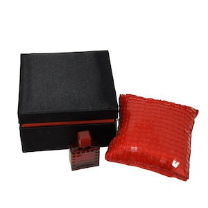 May include: A square black gift box with a red interior trim, a small dark red glass perfume bottle with a geometric textured surface, and a square red pillow covered in small, reflective, square-shaped red sequins.