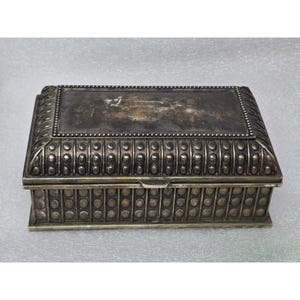 May include: A rectangular metal box with a dark, tarnished finish. The lid features a central flat panel surrounded by a beaded border, while the sides and top edges are decorated with a repeating pattern of raised, vertical oval studs.
