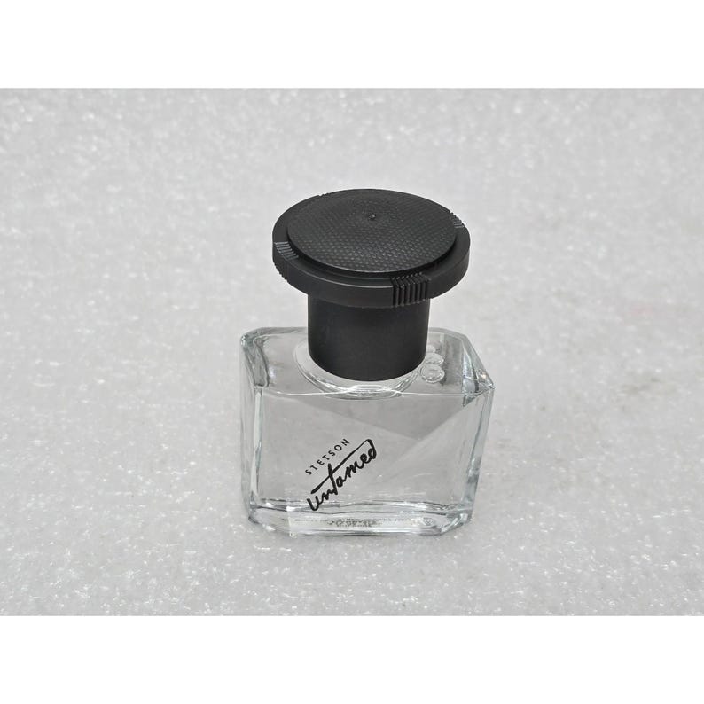 May include: A clear, square glass bottle with a black cap. The bottle has the text "Stetson Untamed" in a cursive font. The cap is round and textured. The bottle is sitting on a white surface.